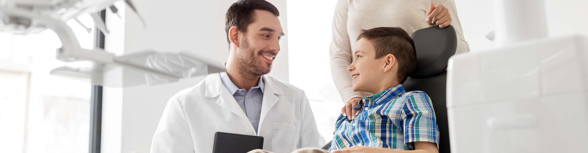seated dentist talking to boy in dentist chair with mom standing behind him