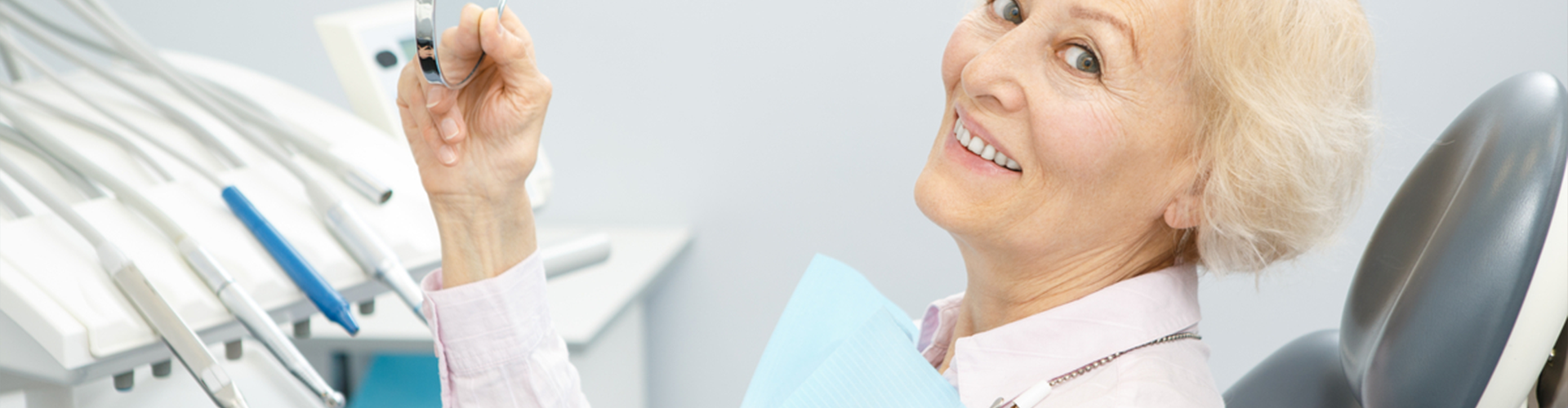Older woman sitting in dentist chair holding a mirror up and smiling 